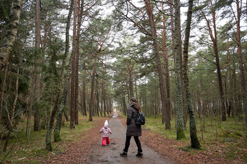 Little girl wear jacket and hat walk is playing at the forest  with mother near baltic sea on cloudy sky in winter time. back view