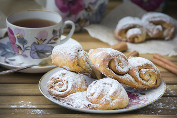 Freshly baked rolls of curd pastry, sprinkled with powdered sugar and a mug of hot tea on a wooden surface, daylight