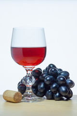 A glass of red wine next to bunch of black grapes and a cork on a wooden table on a white background (concept)