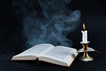 Smoke comes out of old book and candle burning in old candlestick on dark black background