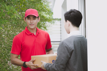 Obraz premium Young logistic career concept. Happy delivery man giving his package to customer at home. Taken in real house. Asian chinese fit man in red polo shirt and jeans with red hat in his early twenties.