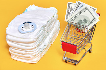 babies diapers with pacifier near shopping trolley cart with money and baby stuff on yellow background