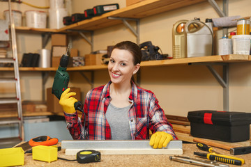 Beautiful caucasian young brown-hair woman in plaid shirt and gray T-shirt working in carpentry workshop at table place, drilling with drill holes in piece of iron and wood while making furniture.