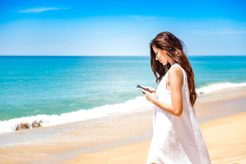 Fototapeta na wymiar Beautiful young single white woman on beach. Using mobile phone calling her friends, wearing white dress. Connected world concept.