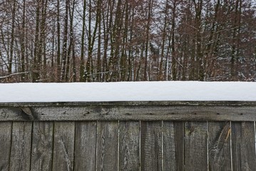 часть серого деревянного забора под снегом у деревьев - obrazy, fototapety, plakaty