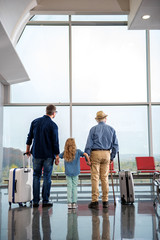 Full length of friendly father, daughter and grandfather are standing near big window at airport while looking through glass. They are holding by hands while carrying suitcases. Focus on their back
