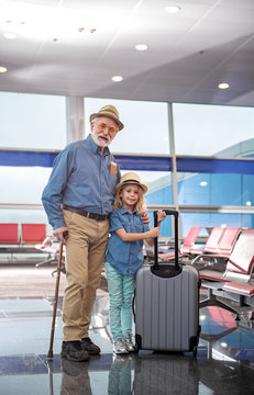 Full length portrait of optimistic gray-haired grandfather and his little granddaughter are standing together at airport lounge. They are looking at camera with smile while girl is holding suitcase