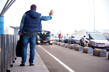 Full length back view of adult man with backpack is standing near road while raising hand. He is trying to catch car. Copy space in the right side. Selective focus