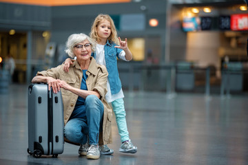 My best friend. Full length portrait of trendy optimistic senior grandmother is squatting and resting on suitcase in airport while her grandchild is hugging her for shoulders. Copy space in right side