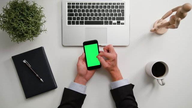 A man's hands holding a phone and touching the green sreen over a white table. View from the top. Close-up shot