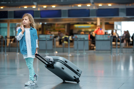 First journey. Full length of stylish cute blond child is standing at airport building and carrying her suitcase with toy bear. She is looking aside pensively while waiting for flight. Copy space