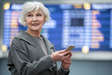 Portrait of joyful charming old woman is standing at international airport against electronic...