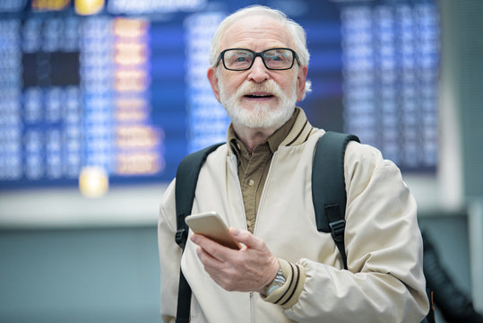 Full of positive emotions. Portrait of cheerful bearded gray-haired man is standing at airport and holding mobile phone while looking aside with smile. Flights departures in background. Copy space