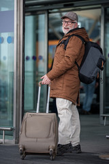 See you. Low angle full length of joyful aged bearded man with backpack is entering to the airport while carrying his suitcase. He is looking at camera with wide smile