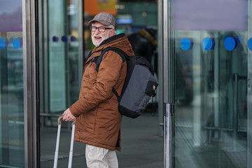 Lets travel. Side view of optimistic senior man with glasses is standing with backpack near entrance to airport. He is holding luggage while looking at camera with joy. Copy space in the right side