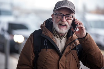 Cheerful news. Portrait of positive gray-haired man with stubble is standing on street while looking at camera with joy. He is having pleasant conversation on modern mobile phone