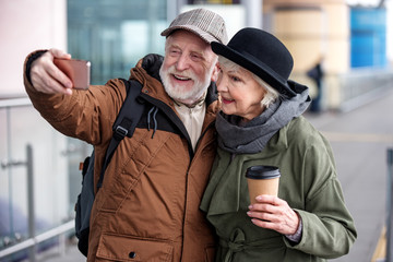 Fototapeta premium For memory. Elegant stylish cheerful old lady with fresh espresso and gray-haired positive man with backpack are taking selfie while standing outdoors. They are hugging with smile