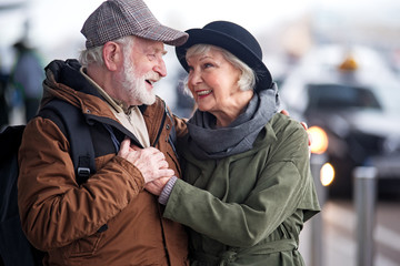 Fototapeta premium My love with you. Waist up of pleasant elderly bearded man with backpack and charming old woman are standing outdoors and hugging. They are looking at each other with fondness