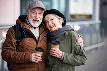 Fototapeta premium Together forever. Waist up portrait of happy aged couple is standing together outdoors and holding coffee. Men is hugging elegant woman and expressing admiration. They are looking at camera with joy
