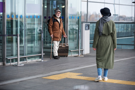 Finally together. Full length of senior man is exiting from airport building while carrying suitcase. He is expressing gladness while seeing stylish woman who is waiting for him. Back view of lady
