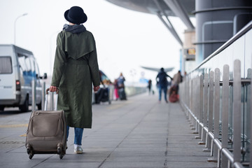 Obraz premium Start of journey. Full length of elegant lady in hat is carrying her suitcase while walking to the airport. Back view. Copy space in the right side