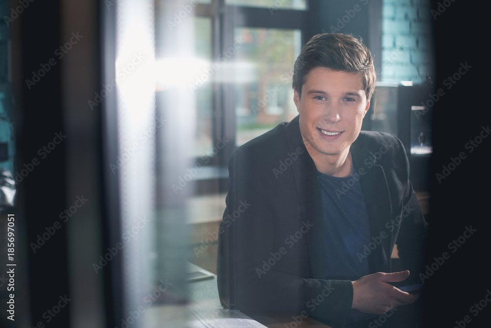 Poster portrait of smiling male holding phone in hand while locating at desk in room. he looking at window.