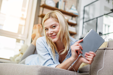 Low angle portrait of blond girl reposing on cozy lounger in apartment. She is holding tablet and...