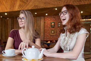 Flirty young girlfriend drinking tea at a table in a cafe with enthusiastic happy faces watching what is happening around. Interested and laughing expression.
