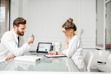 Plakat Senior doctor with young female assistant working on medical documents sitting together at the white office interior