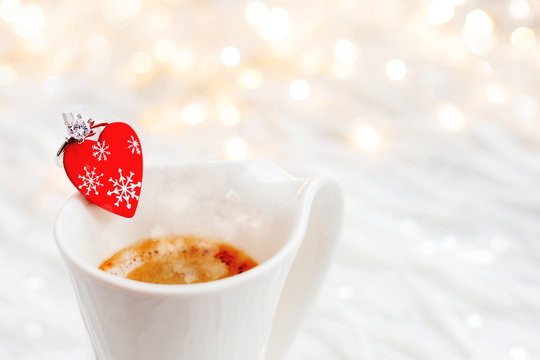 White cup of hot coffee with decorative heart and engagement diamond ring, symbol of love and marriage. Valentine's day background with lights and decorations.