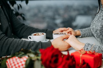 Loving couple celebrating Valentines day holding hands having date at cafe with roses and gift on foreground. Relationships, togetherness, love, romantic concept