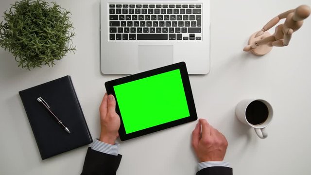 A man's hands holding an i-Pad and touching the green sreen over a white table. View from the top. Close-up shot.