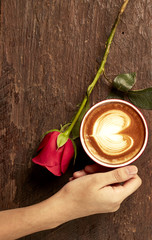 Hand of a young woman holding a red coffee cup on an old wooden desk with roses, conveying love.