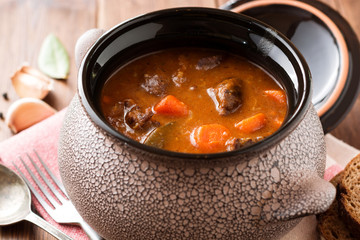 Meat stew with vegetables in ceramic pot on wooden table.