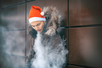 Vape man. Portrait of a handsome young white guy in a Santa Claus hat and winter jacket vaping an electronic cigarette opposite the futuristic urban