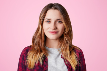 Pleased pleasant looking young female with long straight hair stands against pink background, being in good mood after having date with handsome stylish guy, rejoices starting new relationships