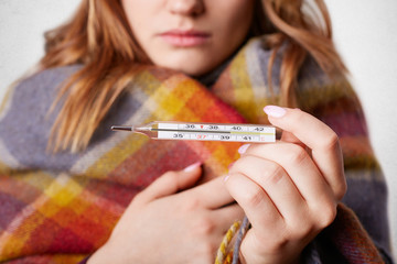 Cropped shot of young female wrapped in warm coverlet, holds termometer which shows high...