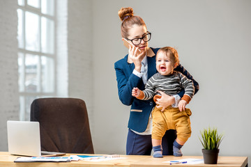 Young multitasking businessmam dressed in the suit talking phone standing with her baby son at the...