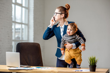 Young multitasking businessmam dressed in the suit talking phone standing with her baby son at the office