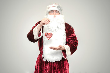 Fototapeta na wymiar Kind Santa Claus holds in hands a red heart shape Christmas toy isolated on gray background.