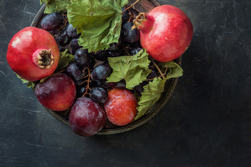 Organic Purple Grapes Red Plums Pomegranates on Vintage Plate. Black Stone Background. Autumn Fall Still Life Harvest Jewish New Year Rosh Hashanah Thanksgiving Top View Copy Space