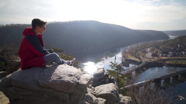 Young man sits on a rock at Maryland Heights while looking at the view of Harpers Ferry National Park and the Potomac and Shenandoah Rivers.