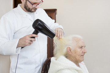 Fototapeta premium Pielęgniarz suszy suszarką włosy starej kobiety siedzącej na krześle.
