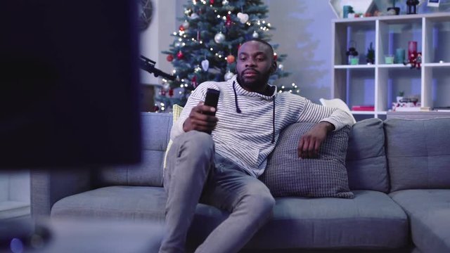 Young handsome African man in casual clothes sitting on sofa and viewing television. Disgruntled guy switching TV channels. Indoors.