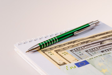 American one hundred dollar bills. Euro banknotes. A ballpoint pen and a notebook next to the bills. Business and finance. Money. White background.