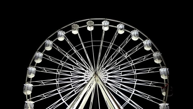 Front view of illuminated white ferris wheel carnival ride revolving isolated against plain black sky background at night