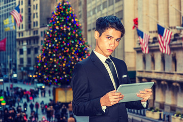 Asian American college student studying in New York. Wearing black suit, necktie, holding small computer, a businessman standing on street in winter holiday season, reading. Filtered look..