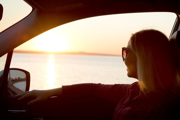 Fototapeta premium A young woman looks out the car window at the sunset on the sea. 