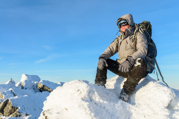 Fototapeta na wymiar Hiker with backpack sitting and having rest on the top of a snow-covered rock over the winter mountains