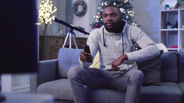 Handsome guy sitting on couch in nice living room and viewing football match. Smiling African man looking directly at screen on background of Christmas tree.